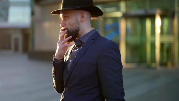 Bearded Man with Stylish Moustache and Elegant Hat is Smoking in City Street in Sunset Time