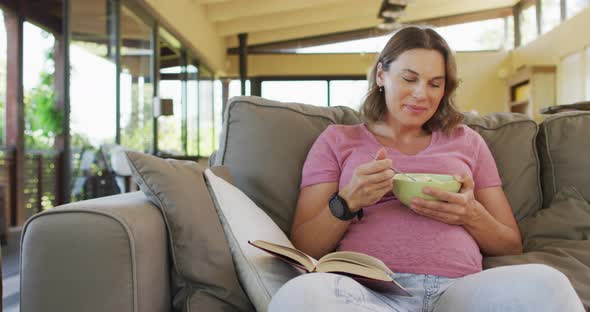 Relaxed caucasian pregnant woman lying on sofa, eating and reading book