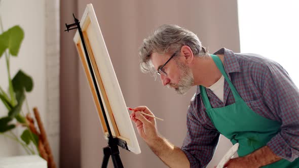 Man artist in glasses and an apron paints on canvas with brush with paints at living room, side view