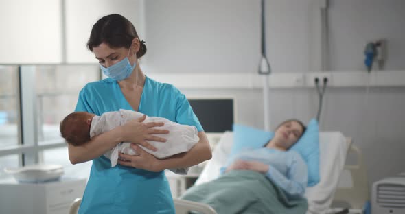 Nurse in Medical Mask Holding Newborn Baby with Mother Resting in Bed on Background