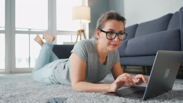 Woman with Glasses Is Lying on the Floor and Working on a Laptop