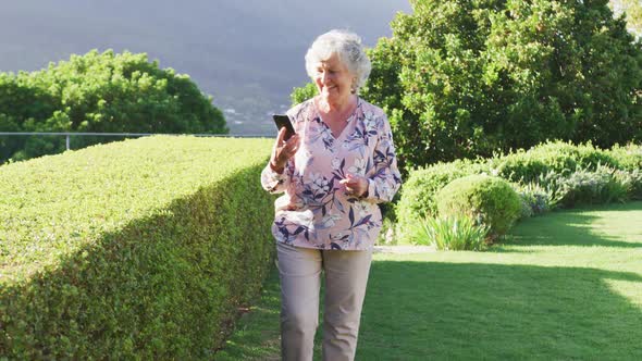 Caucasian senior woman smiling while talking on smartphone while walking in the garden