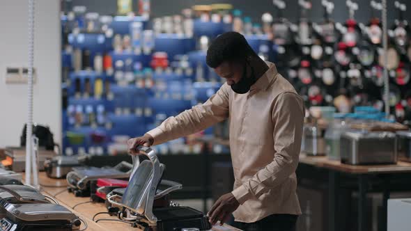 Young Black Man with Face Mask is Shopping in Home Appliances Store Viewing Samples of Electric