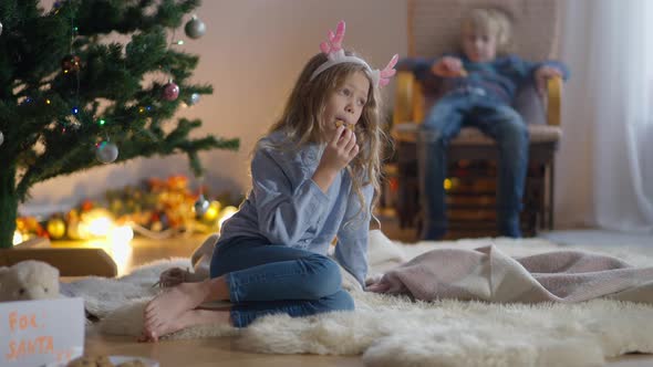 Wide Shot Portrait Charming Little Thoughtful Girl Eating Christmas Cookie with Boy in Rocking Chair