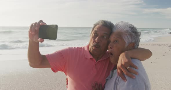 Happy hispanic senior couple making funny faces, taking selfie on beach at sunset