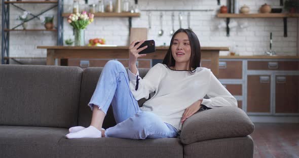 Young Asian Teenager Woman Lying in Couch Using Smartphone Video Conference with Friends While Lying