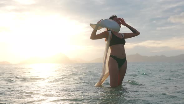 Woman In Bikini Holding Sun Hat Standing In Sea At Sunset