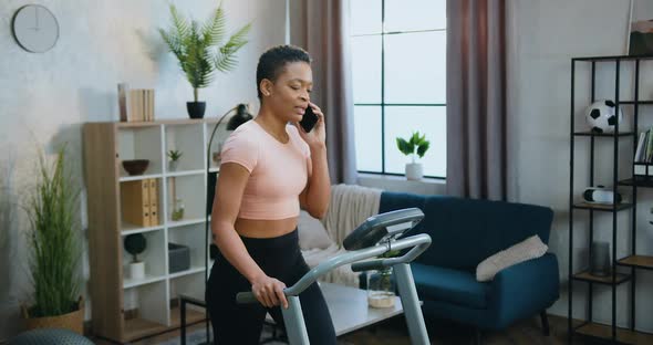 Woman in Training Clothes Walking on Treadmill During Home Morning Workout and Talking on Phone