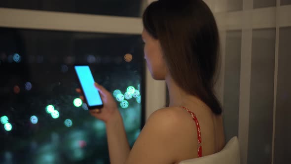 girl holds phone and looks at it at window against background of night city.