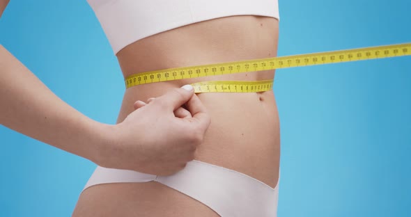 Young Slender Lady in Underwear Measuring Her Waist with Meter Side View Blue Studio Background