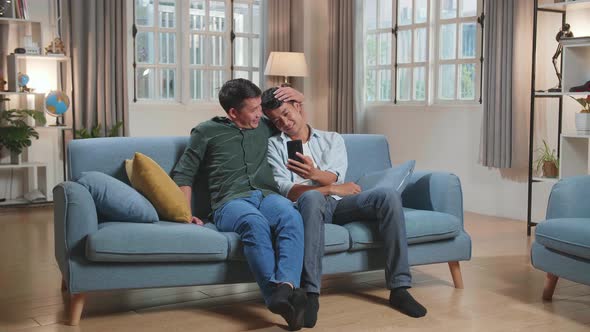 Asian Gentle Gay Couple Using Mobile Phone While Sitting On A Couch In Apartment