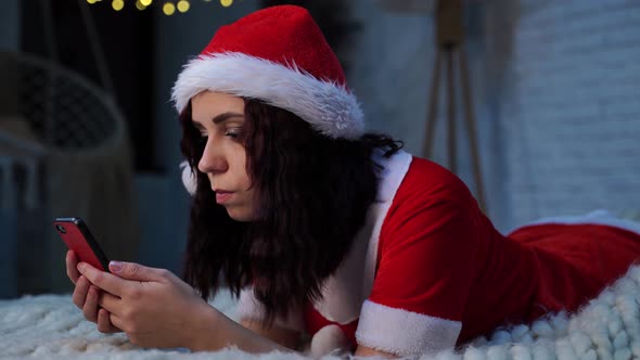 Young Woman in Santa Claus Costume Browses Smartphone Lying on Bed