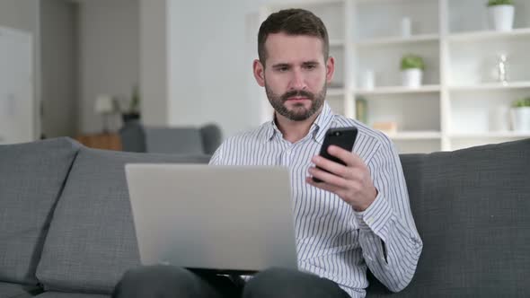 Young Man with Laptop Talking on Smartphone at Home