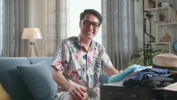Asian Man Packing Clothes In Suitcase And Warmly Smile To Camera At Home, Preparing For Vacation