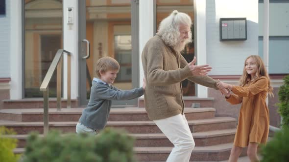 Side View of Cute Girl Pulling Hand of Old Man As Cheerful Boy Pushing His Back. Joyful