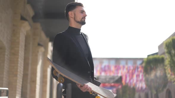 Panning Shot of Elegant Caucasian Businessman with Skateboard Standing in Sunrays Outdoors