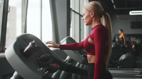 Beautiful woman of athletic build turn on treadmill in gym and starts jogging, side view.