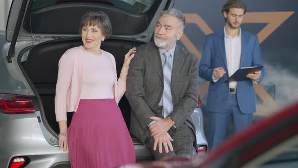 Smiling Senior Couple of Retirees Choosing New Automobile in Car Dealership with Dealer Standing at