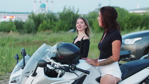 Two Women Friends Sitting on Motorcycles Near the Water of Evening City and Talking