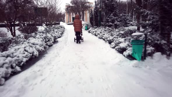 Handheld Firstperson View  Mom with a Baby Carriage Walking Along a Snowcovered Frosty Alley