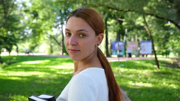 Young Woman Turns and Looks at the Camera on the Background of the Park 