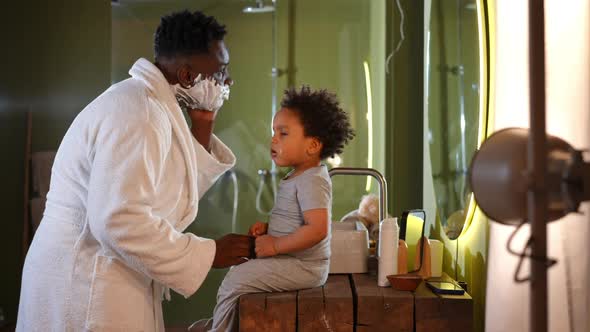 Playful Young Father Applying Shave Foam Talking to Cute Toddler Son Sitting in Bathroom