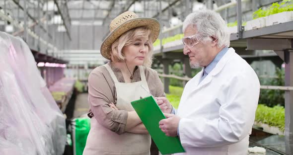 Portrait of Worried Caucasian Female Agronomist Giving Task To Male Senior Scientist in Glasshouse