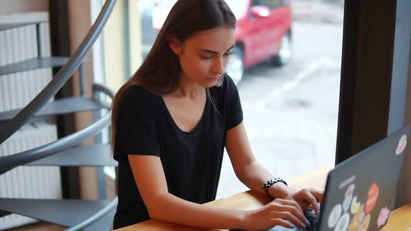 Young Woman Sitting in the Coffee Shop By the Window with Her Laptop and Thinking What to Write or