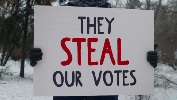 Protestor standing with banner with black and red writing They Steal Our Votes.