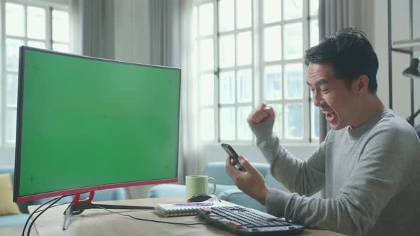 Successful Asian Man With Green Screen Computer Looking At Mobile Phone While Working At Home