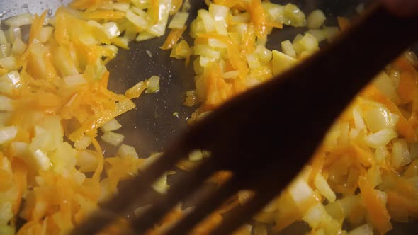 In a black skillet, fry carrots and onions. A wooden spatula stirs them. Slow motion