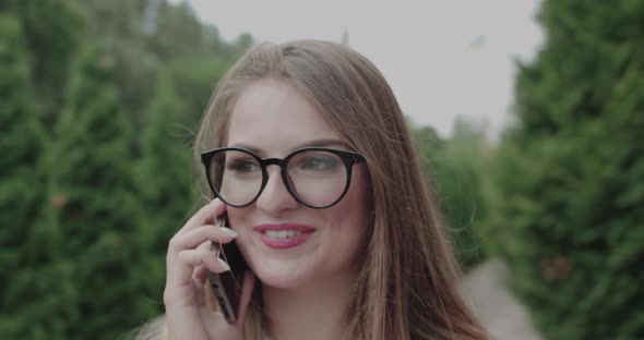 Cheerful Young Girl in Glasses Speaking on a Phone with Smile on Face Outdoors