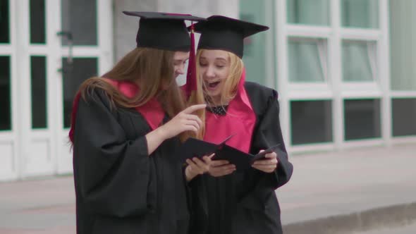 Two Beautiful Female Caucasian Graduates Look at Diploma and Rejoice. Outside