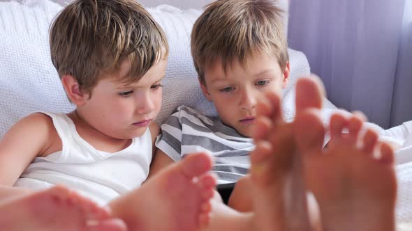 Children get bored. Two boys holding smartphone, tablet sitting on chair, focus on children feet.