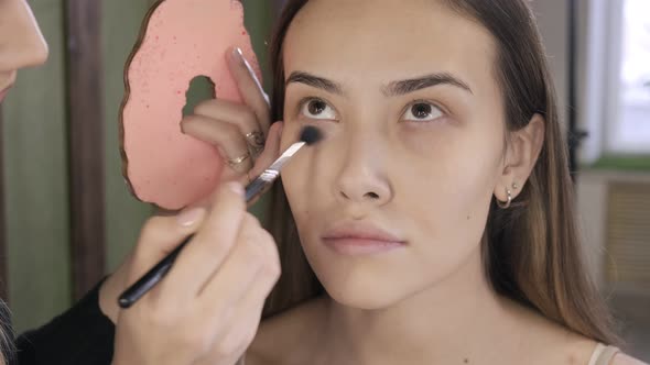 Make Up Artist Doing Makeup for Brunette Asian Woman with Brush in Beauty Salon