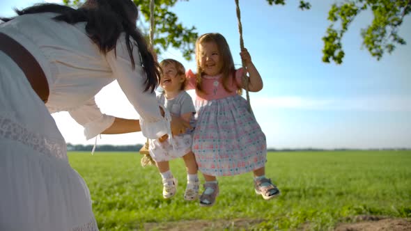 Happy Woman in a White Dress Swings Her Daughters on a Swing.