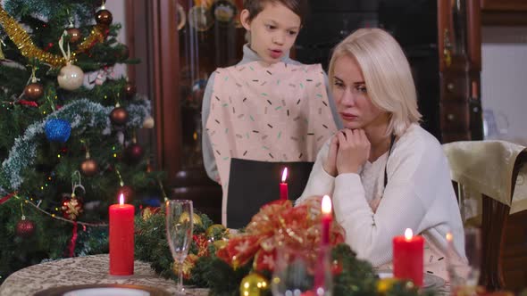 Cute Caucasian Boy Cheering Up Sad Woman Sitting at Decorated Christmas Table at Home