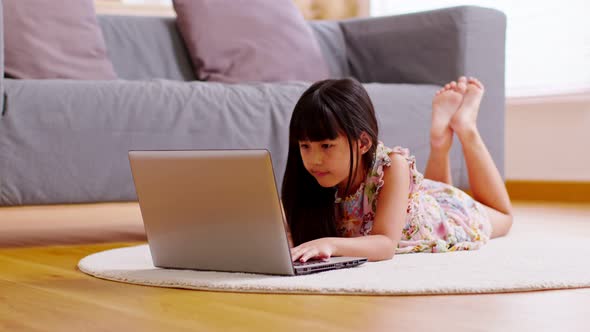 Cute little girl uses laptop while prone at the sofa in the living room. Child surfing the internet