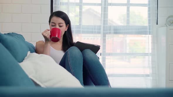 Asian woman using tablet holding a warm cup of coffee or tea while lying on the sofa.