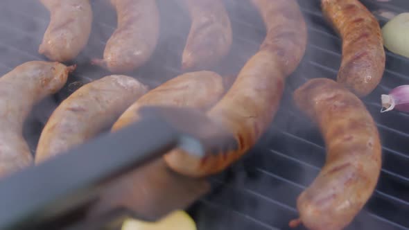 Hand with the Help of Metal Tongs Turns the Simmering Sausages on a Black Barbecue Grill