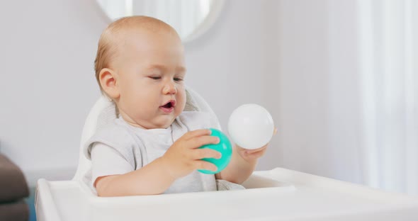 Closeup of Young Baby in White High Baby Chair Who Holds Two Plastic Balls Green and White in Both