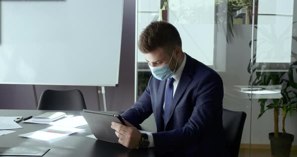 Portrait Caucasian Man in Medical Mask Sitting at Work Table Looking at Screen of Tablet Computer