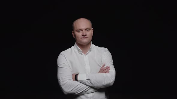 A bald man in a white shirt crosses his arms and poses for the camera on a black background.