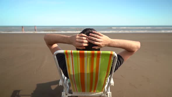 Man Sits On A Chair And Put His Hands At The Back Of His Head, Relaxing At Sandy Beach In Monte Herm