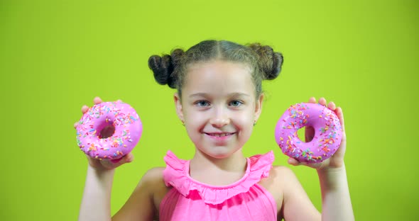 Cute Little Girl Holding Donuts with Icing in Her Hands, Smiling, Looking at the Camera and Bringing