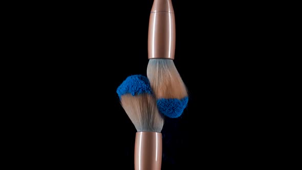 Two Makeup Brushes Collide and Cause a Swirl of Blue Powder Particles Against a Black Background