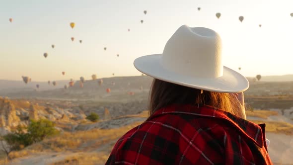 Woman In Hat Walking In Cappadocia. Colorful Hot Air Balloons Fly On Background