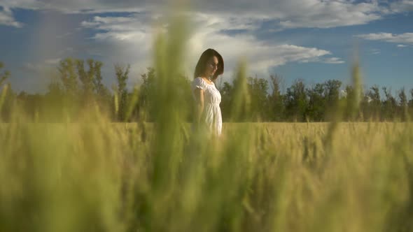A Young Woman in a White Dress Walks on a Green Wheat Field. View of the Girl Through the Spikelets.