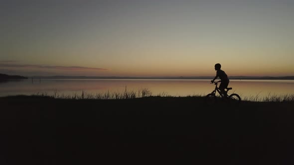 Silhouette of happy young boy riding his bike on shore next to beautiful calm lake reflecting vibran