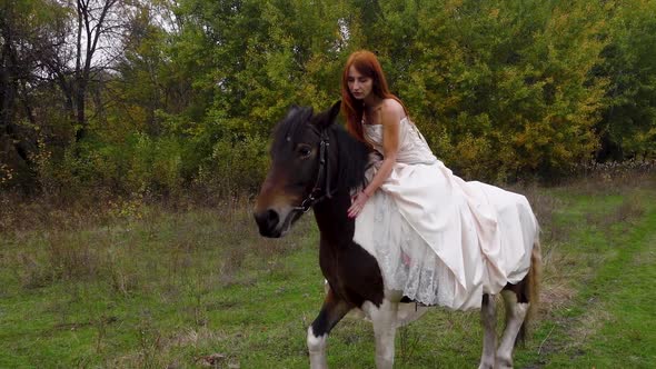 Redheaded Girl in Beige Dress Is Riding Horse Near Wood